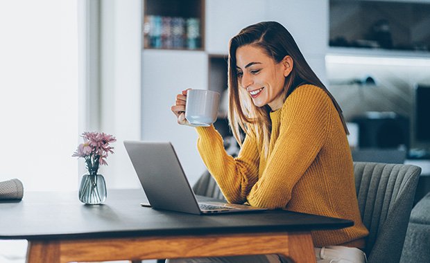 Woman wearing yellow sweater sits at kitchen table using laptop