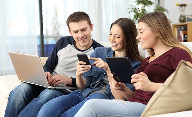 Smiling young man and two young women sit on couch looking at phone held by woman in the middle