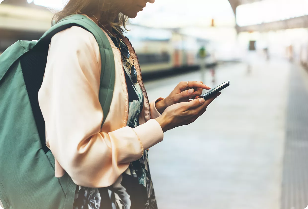 A person standing on a train platform checks their smartphone while wearing a backpack.
