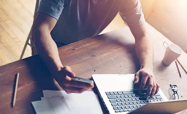Hand holds a smartphone over a pile of papers on a desk while the other hand rests on a laptop keyboard