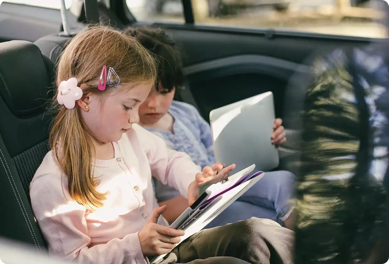 Two young children sitting in the back seat of a car using tablets, focused on their screens.