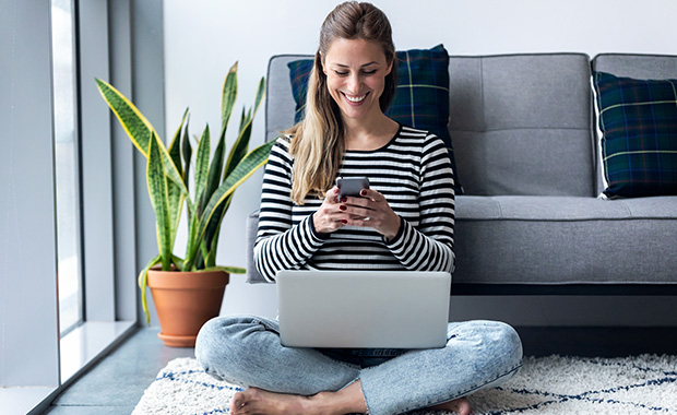 Young woman using her smartphone while working on her laptop sitting on the floor at home