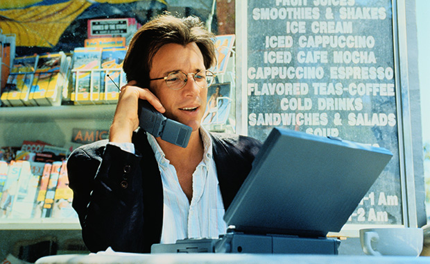 Man sits in café working on laptop and speaking on cell phone. The devices date the scene to the late 1990s