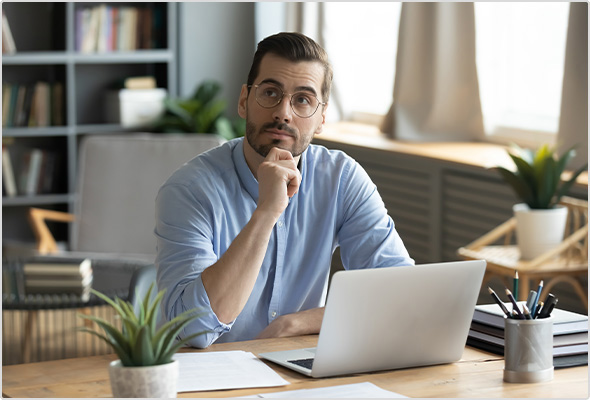 Man sits at desk in front of laptop with hand on chin