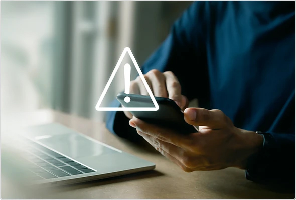 A person sitting at a desk using a smartphone next to an open laptop with a warning icon overlayed.