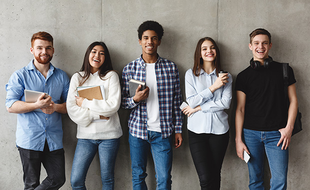 Group of students leaning on a gray wall and smiling at the camera.