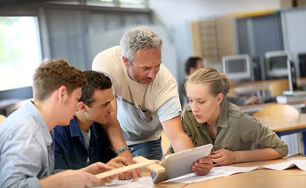 A teacher assists a group of students in a classroom as they collaborate using a tablet, blueprints, and a wooden model.